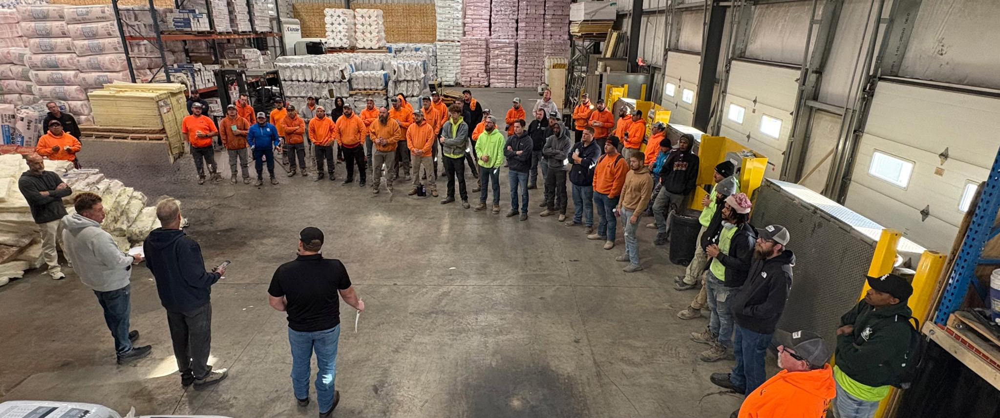 Workers in an orange-vest safety briefing forming a circle inside a busy warehouse.