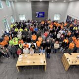 Overhead shot of a large group of employees posed for a group photo in a modern lobby, many wearing orange safety shirts or high-visibility vests.