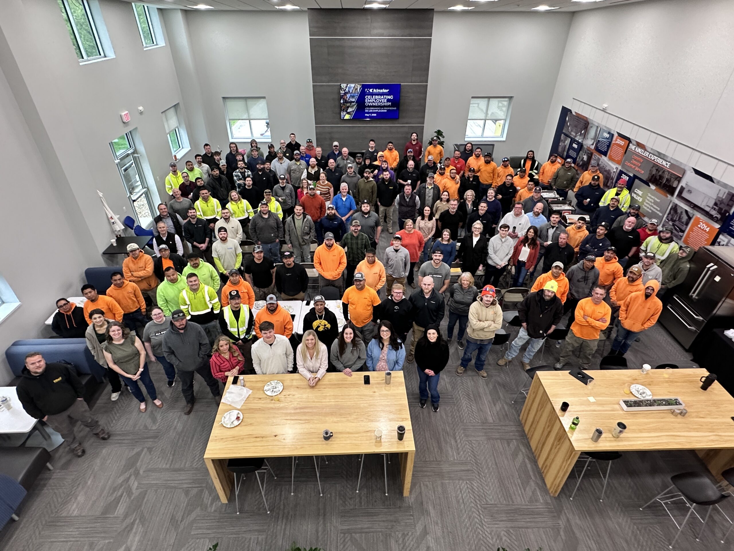 Overhead shot of a large group of employees posed for a group photo in a modern lobby, many wearing orange safety shirts or high-visibility vests.