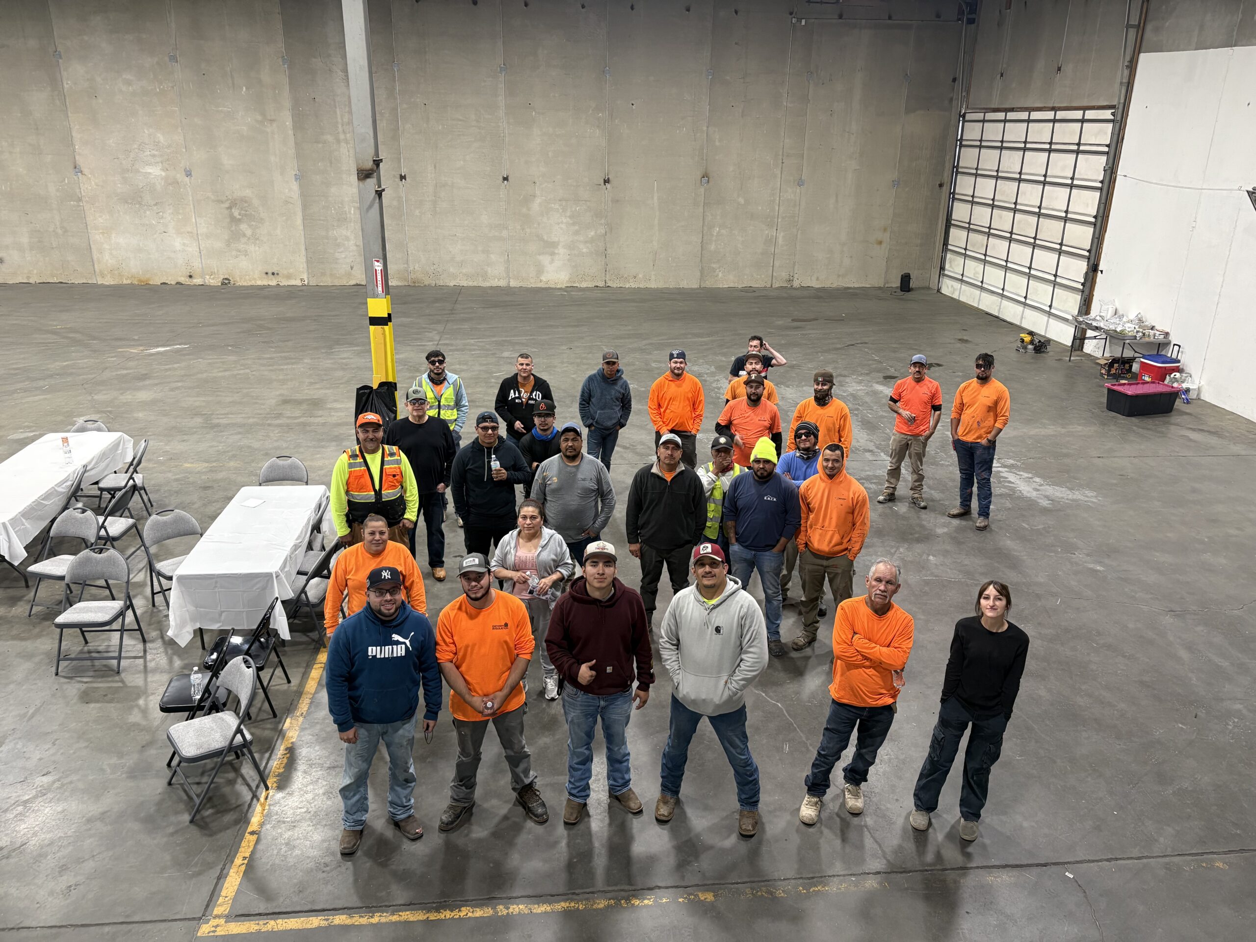 Group of workers in a large, empty warehouse, many wearing orange shirts and safety vests, posing for a photo.