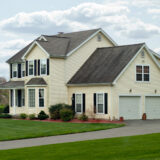 Two-story beige house with multiple gabled roofs, black shutters, and a two-car garage, set on a green lawn.